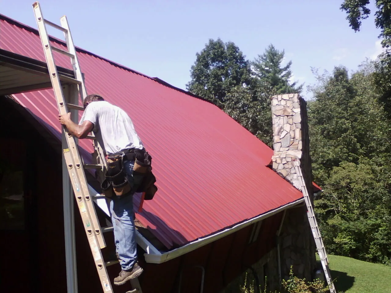 Roofer on a ladder beside a red metal roof and stone chimney — Soffit & Fascia Repair by Roofing Brothers in Oakridge